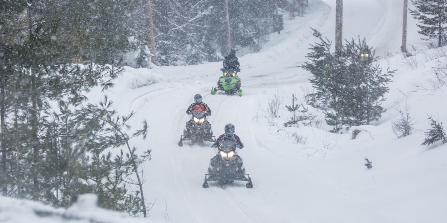 A snowy landscape with a group of snowmobiles on a trail surrounded by trees.