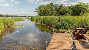 Een houten steiger met stoelen en een tafel, met uitzicht op een rustige rivier langs begroeide oevers.