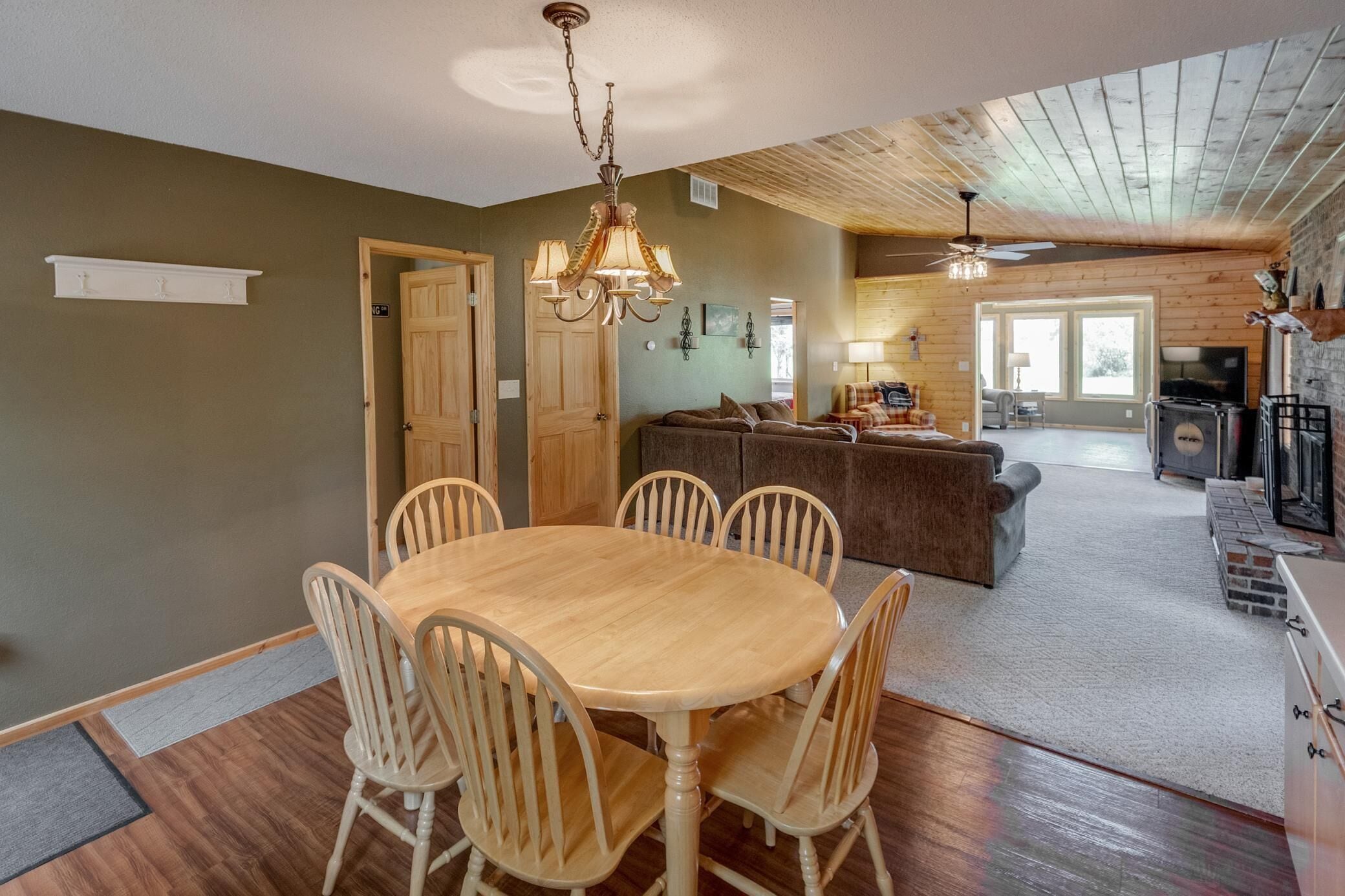 A dining area with a wooden table and chairs, a chandelier, and a kitchen in the background.
