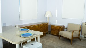 Ein kleiner, gut beleuchteter Raum mit einer Küchenzeile, einem Esstisch mit Tellern, einer Lampe, einem Stuhl und einem Schrank.