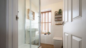 A bathroom with a glass shower enclosure, a white toilet, a window with blinds, and a shelf with plants. - 3 Victoria House, St Ives – Sleeps 4 – Sea Views (St Ives)