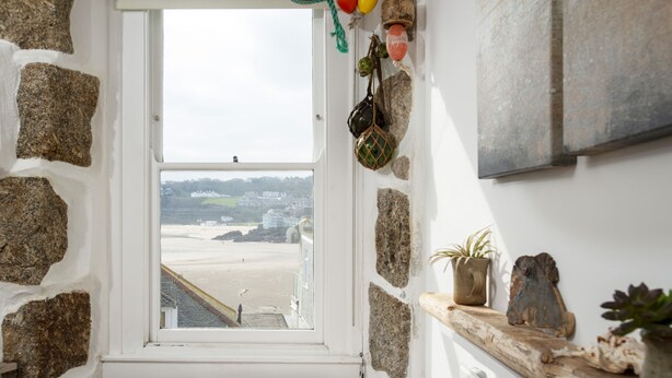 A stone-walled room with a bench, a window overlooking a beach, and a shelf with decorative items. - 3 Victoria House, St Ives – Sleeps 4 – Sea Views (St Ives)