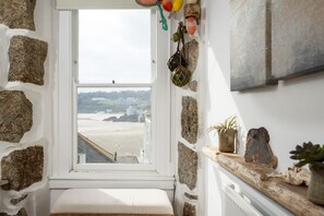 A stone-walled room with a bench, a window overlooking a beach, and a shelf with decorative items.