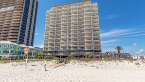 Ein Strandhotel mit mehreren Balkonen, ein sandiger Strand im Vordergrund und ein klarer, blauer Himmel.