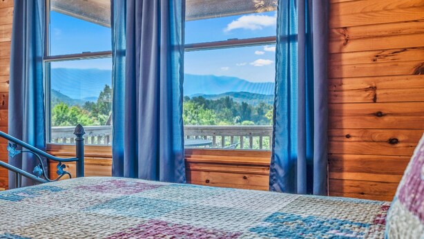 Ein Schlafzimmer mit großem Fenster, Holzboden, einem Bett mit gemustertem Überwurf und Blick auf einen Balkon und Berge.