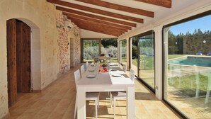 A dining area with a white table set for six, wooden beams on the ceiling, and a view of a swimming pool outside.