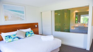 A neatly made bed with white linens and colorful pillows, a wooden headboard, a large window with a view of the beach, and a modern bathroom with a large mirror and sink.