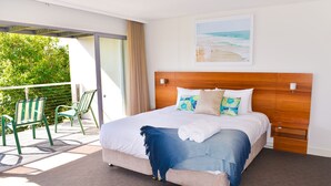 A bedroom with a large bed, a wooden headboard, a balcony with chairs, and a beach view.