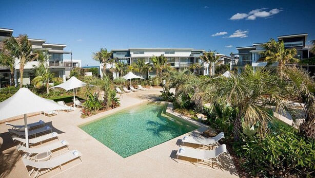 A resort pool area with multiple lounge chairs and umbrellas, surrounded by palm trees and modern buildings.