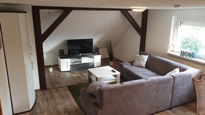 A living room with a grey sofa, white coffee table, and a TV stand with a flat-screen TV. There is a window with a view of greenery outside. - HässelHof - Holiday apartment in the center of Bad Liebenwerda (Bad Liebenwerda)