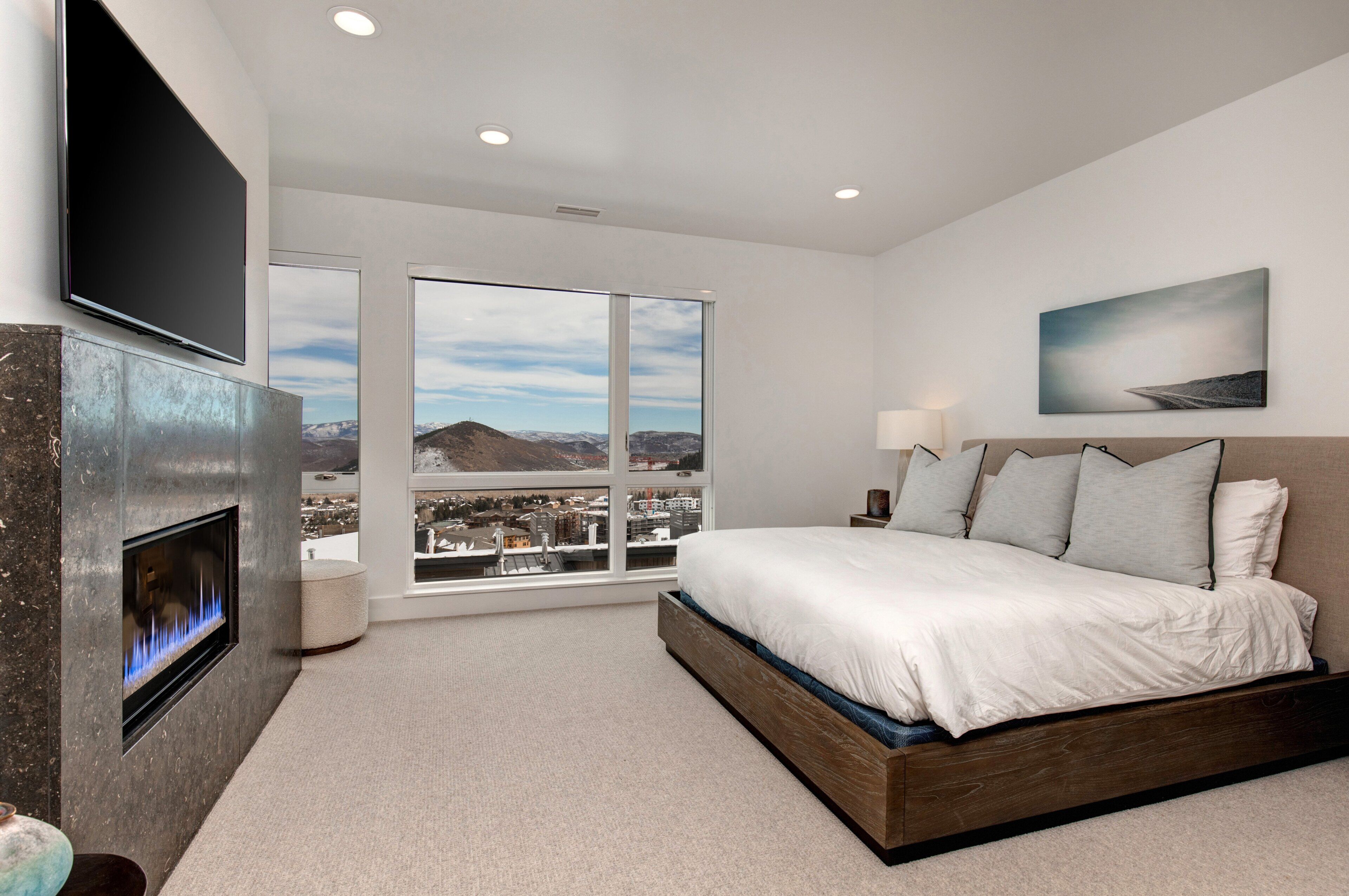 A bedroom with a large bed, a fireplace, a TV, and a city view through large windows.