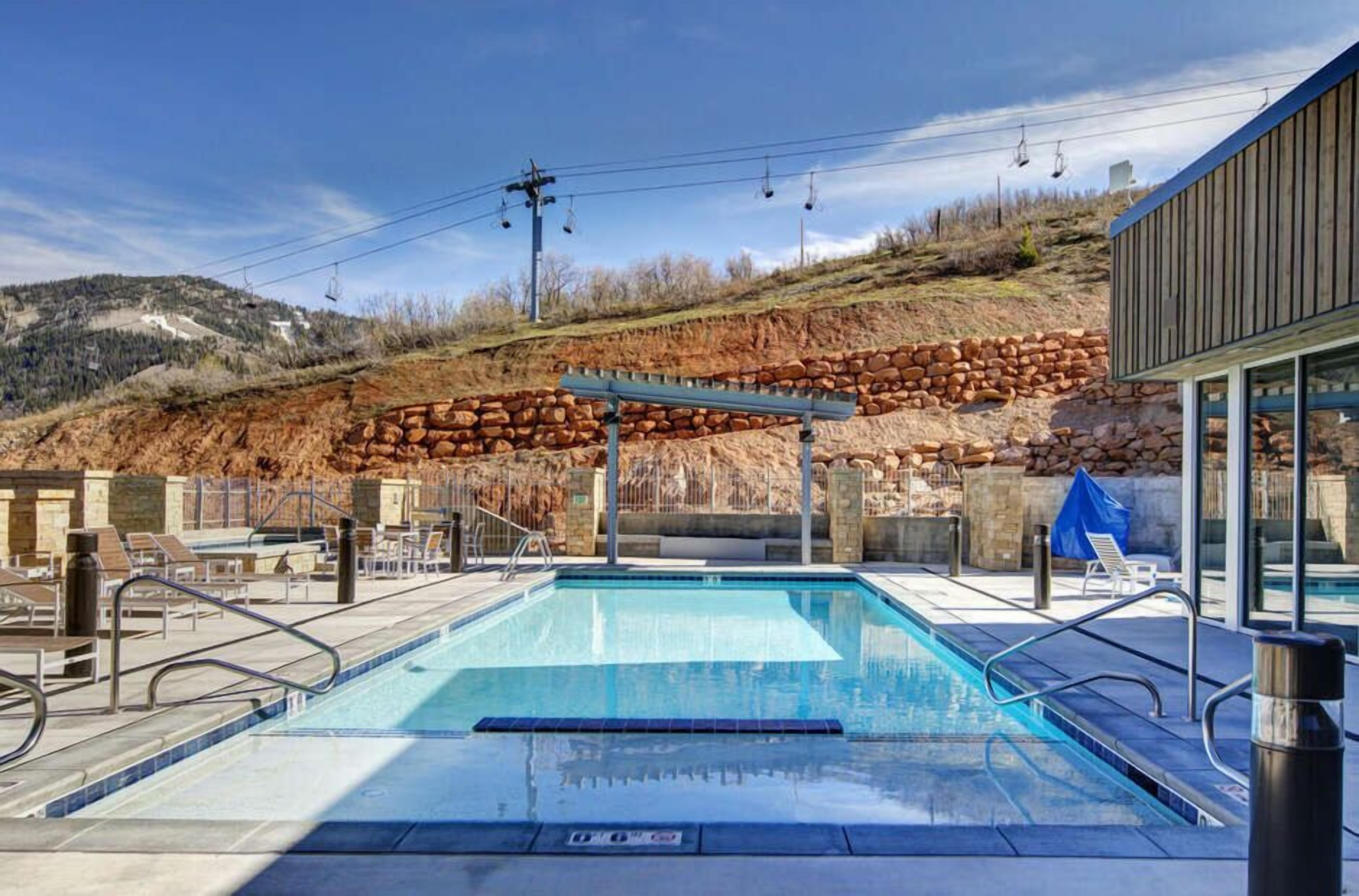 Una piscina con un muro de piedra de fondo, sillas de descanso y un telesilla al fondo.