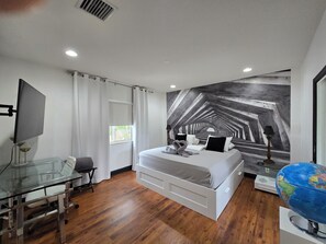 A modern hotel room with a bed, a desk, a chair, a globe, and a large abstract mural on the wall.
