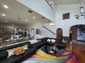 A spacious living area with a large black sectional sofa, a glass dining table set for six, a colorful rug, and a kitchen in the background.