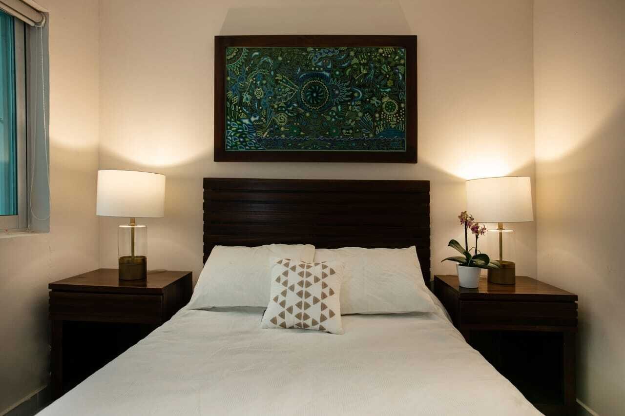Una cama bien hecha con dos almohadas, una obra de arte enmarcada encima y dos lámparas de noche sobre mesitas de madera.