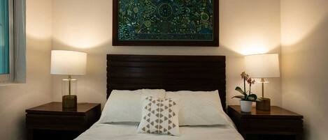 Una cama bien hecha con dos almohadas, una obra de arte enmarcada encima y dos lámparas de noche sobre mesitas de madera.