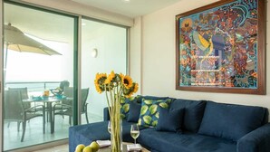 Ein Wohnzimmer mit einem blauen Sofa, einer Glastür, die auf einen Balkon führt, einem Tisch mit einer Vase voller Sonnenblumen und einem farbenfrohen Gemälde an der Wand.