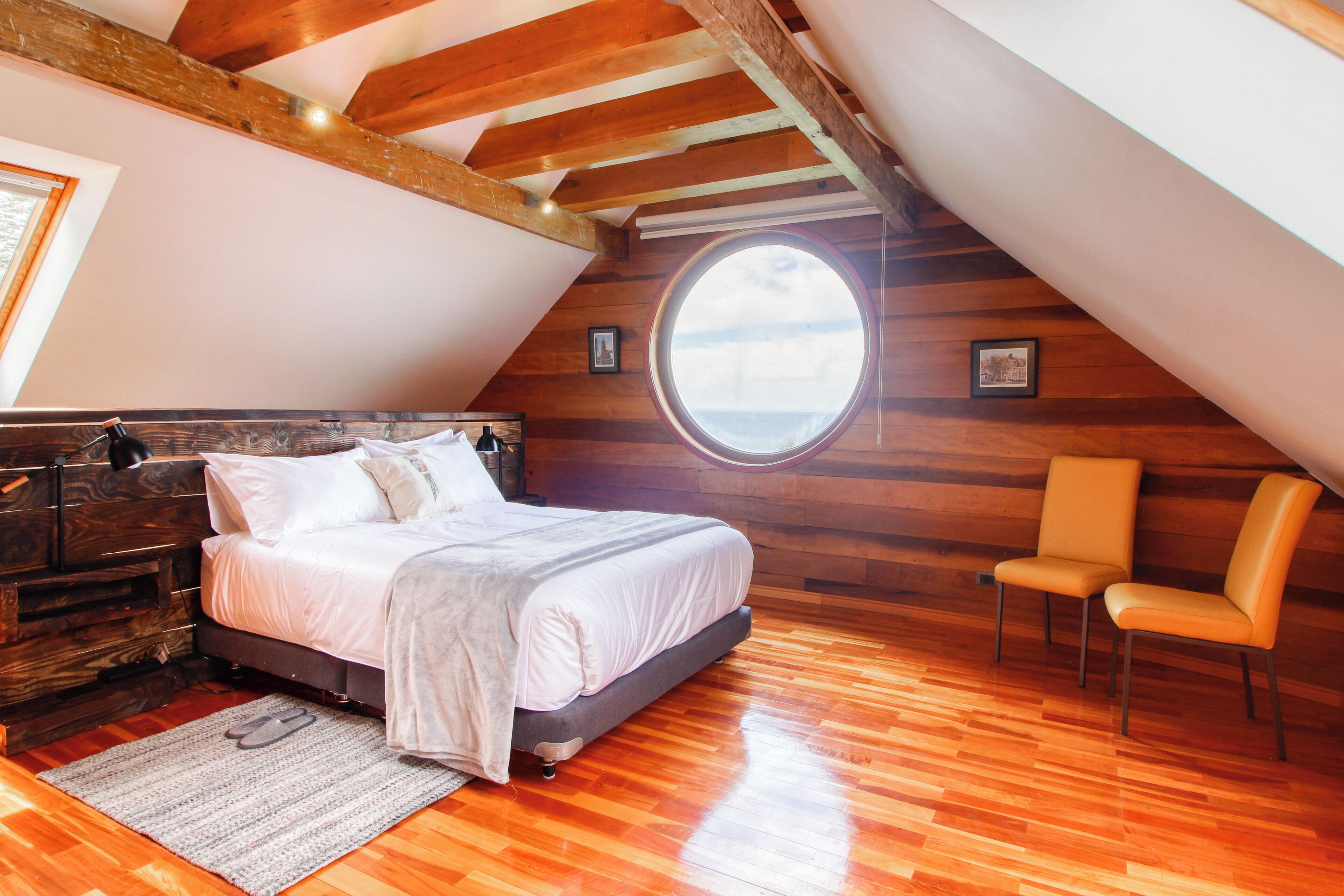 A bedroom with a wooden ceiling, a large round window, a bed, a chair, and a dresser.