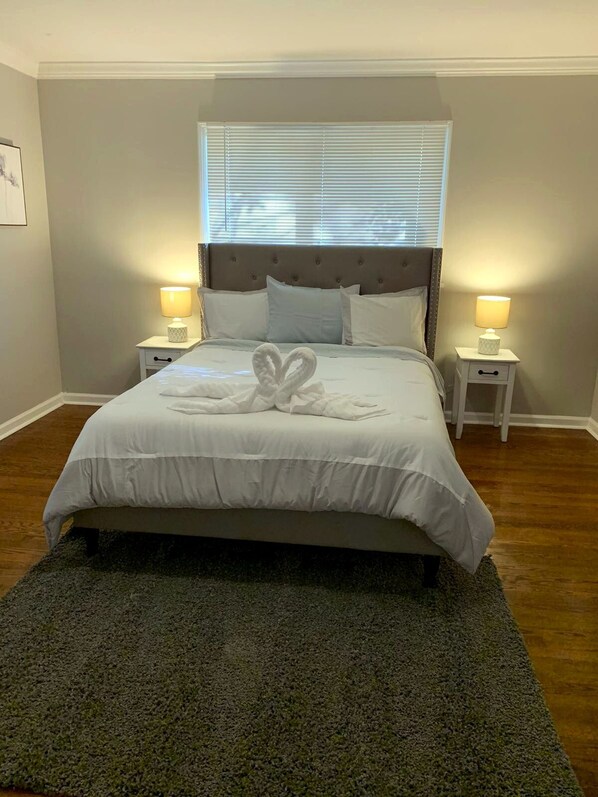 A neatly made bed with a heart-shaped towel arrangement, two bedside lamps, and a window with blinds.