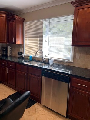 A kitchen with wooden cabinets, a stainless steel sink, a dishwasher, and a window with blinds.