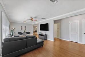 A spacious living room with a grey sectional sofa, a flat-screen TV mounted on the wall, and a ceiling fan.