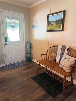 A wooden bench with a cushion, a door with a doormat, a framed picture on the wall, and a small plant on the floor.