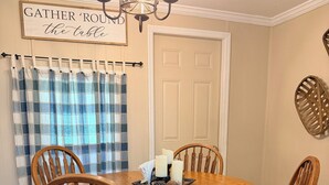 A dining area with a round wooden table, four wooden chairs, a chandelier, a sign above the table, and a door in the background. - Rustic Countryside Cottage (Cleveland)