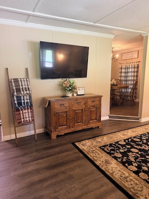 Une pièce avec un meuble en bois, un téléviseur à écran plat fixé au mur, une petite table avec un vase et un panneau indiquant la présence d’une connexion Wi-Fi, et un tapis à motifs sur le sol.