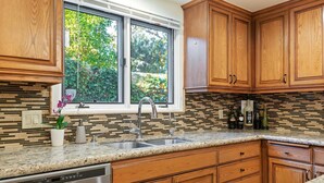 A kitchen with wooden cabinets, a granite countertop, a stainless steel dishwasher, and a window with a view of greenery. - Enjoy the Beauty of Downtown Santa Barbara Walk Everywhere! (Santa Barbara)