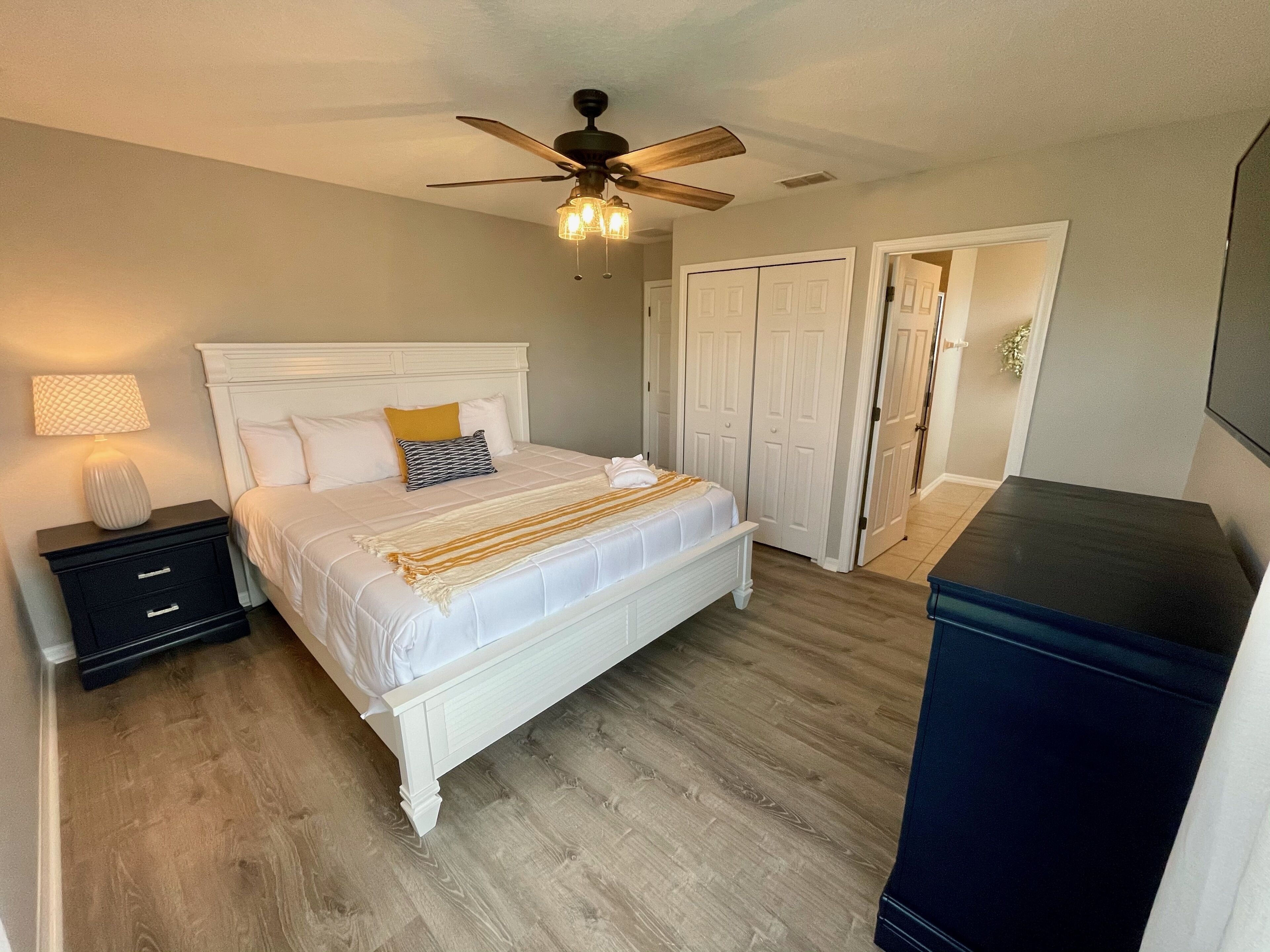 A bedroom with a white bed, a nightstand, a lamp, a ceiling fan, and a wardrobe.