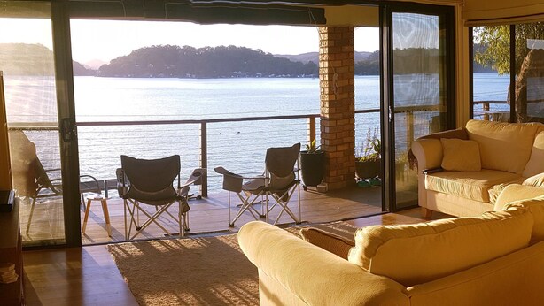 A room with a large glass door leading to a balcony overlooking a body of water. Inside, there is a beige sofa and two folding chairs. - Waterfront with private jetty and sunset views. Boat access only. Pet friendly. (little wobby)