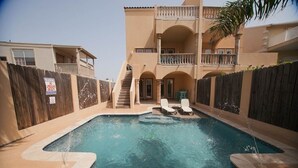 Una piscina con due sdraio, un recinto in legno e un edificio a due piani con balconi.