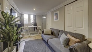 Een woonkamer met een grijze bank, decoratieve kussens, een eettafel met stoelen, een potplant en een groot raam met gordijnen.