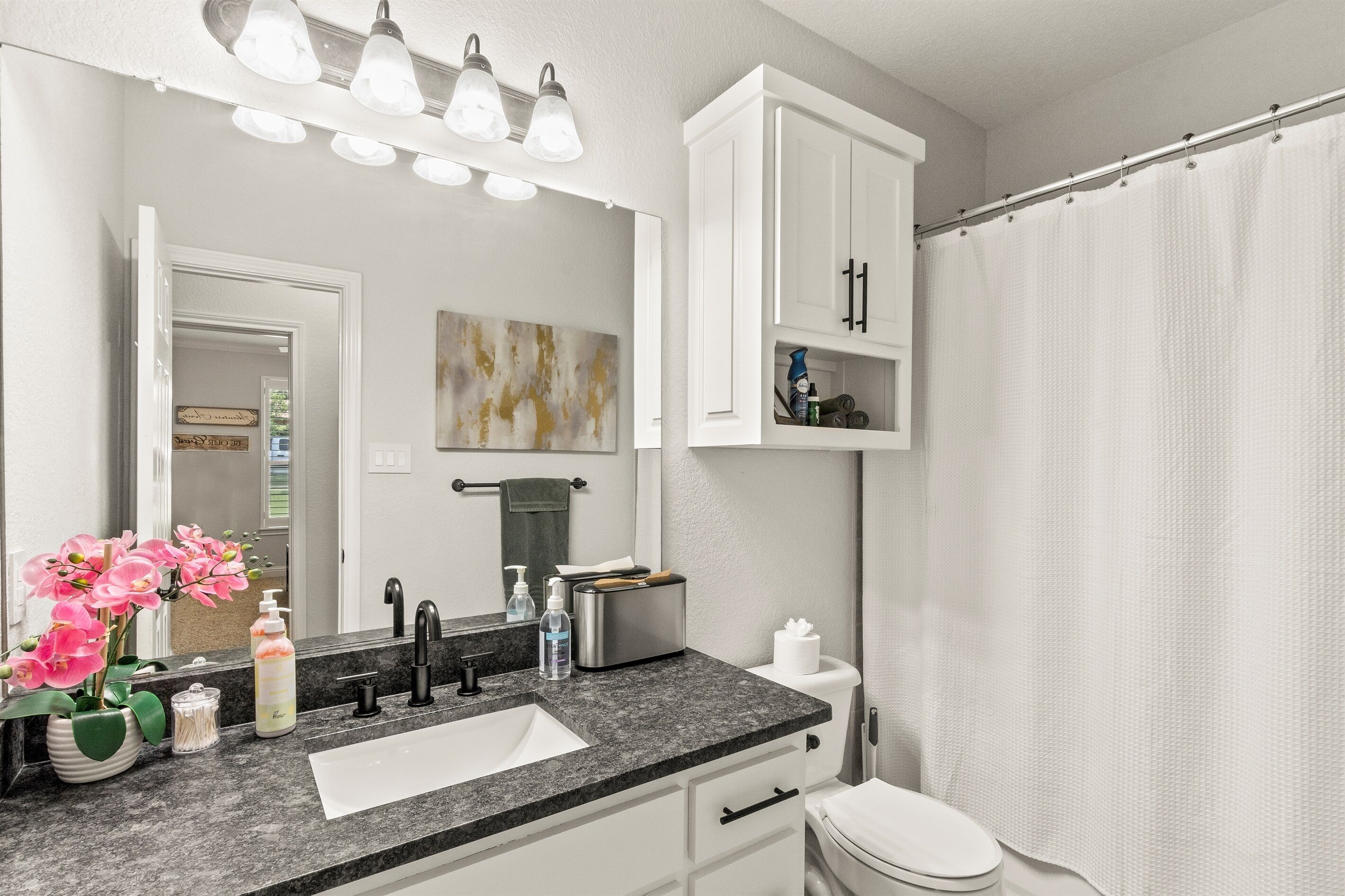 A bathroom with a sink, mirror, and toilet. There are cabinets above the sink and a shower curtain.