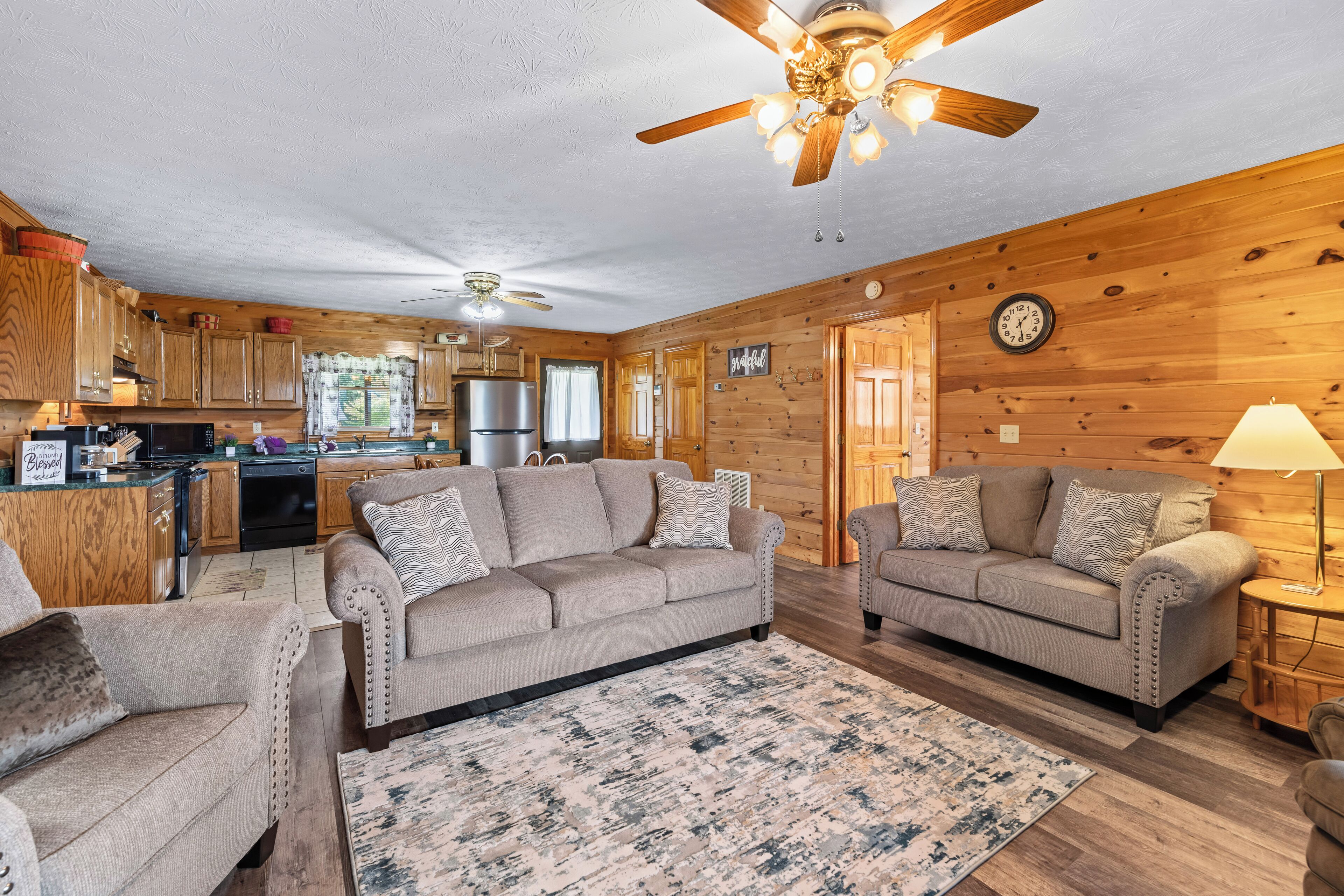 A cozy cabin-style interior with wooden walls, a kitchen with modern appliances, two sofas, and a ceiling fan.