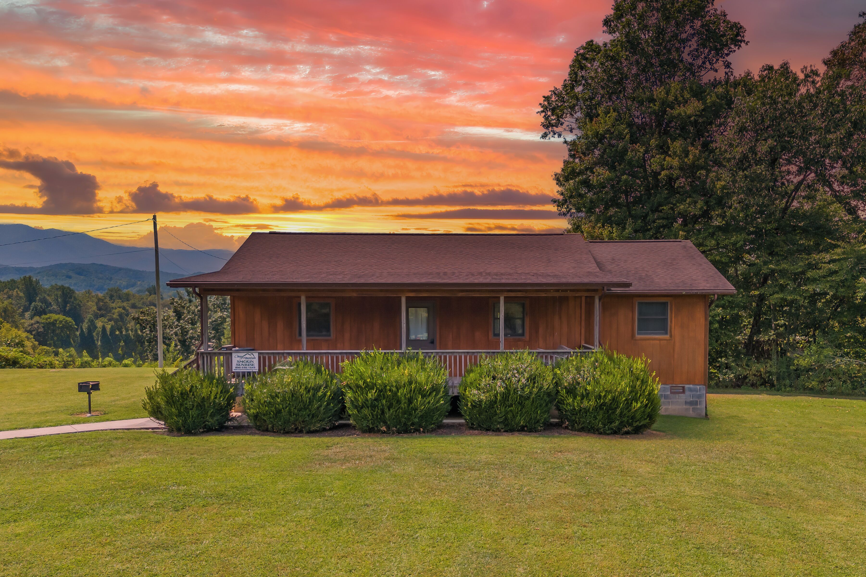 A two-story wooden cabin with a porch, surrounded by well-maintained grass and bushes, set against a backdrop of a sunset sky.