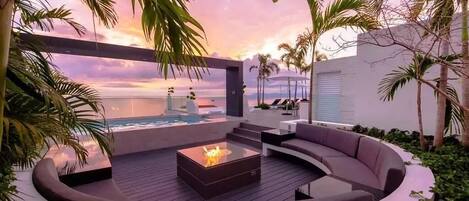 Un espacio circular al aire libre con una chimenea, rodeado de abundante vegetación y vistas a una piscina y al océano.