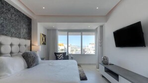 A modern bedroom with a large bed, a TV mounted on the wall, and a view of the city through large windows.