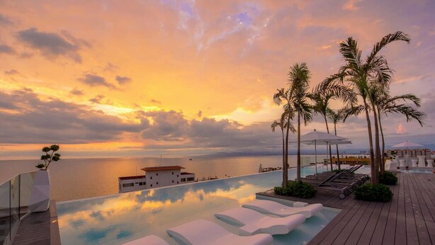An infinity pool with lounge chairs, palm trees, and a view of the sea at sunset.