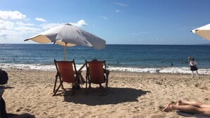 A beach with two chairs under a large umbrella, a person lying on the sand, and the ocean in the background.