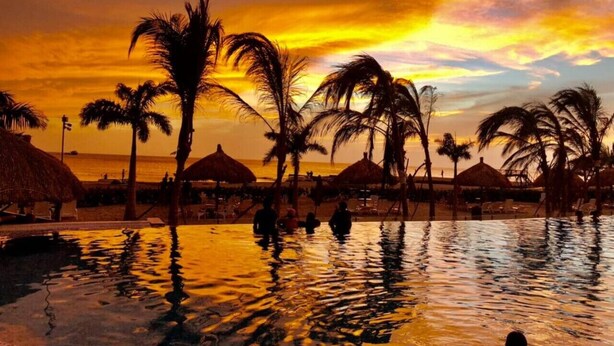 A pool area with palm trees and thatched umbrellas, surrounded by a beach setting. - Beachfront Family Escape | 3BR Samaria Club! (Santa Marta)