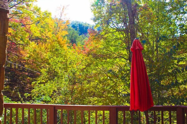 A wooden deck with a red umbrella, surrounded by trees with autumn foliage. - Just 10 minutes from GSM National Park, 2 BR Log Cabin with Hot Tub & Firepit (Pigeon Forge)