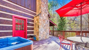 A wooden cabin with a stone chimney, a red door, a hot tub, and a covered deck with red chairs and a table. - Just 10 minutes from GSM National Park, 2 BR Log Cabin with Hot Tub & Firepit (Pigeon Forge)
