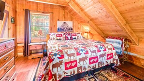 A cozy attic bedroom with a wooden ceiling, a large bed with a quilt featuring animal prints, a window with a view of trees, and a wooden dresser. - Just 10 minutes from GSM National Park, 2 BR Log Cabin with Hot Tub & Firepit (Pigeon Forge)