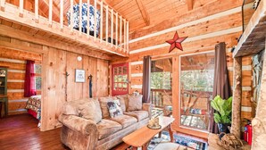 A cozy cabin interior with a wooden floor, a sofa, a table, and a balcony with a red star decoration. - Just 10 minutes from GSM National Park, 2 BR Log Cabin with Hot Tub & Firepit (Pigeon Forge)