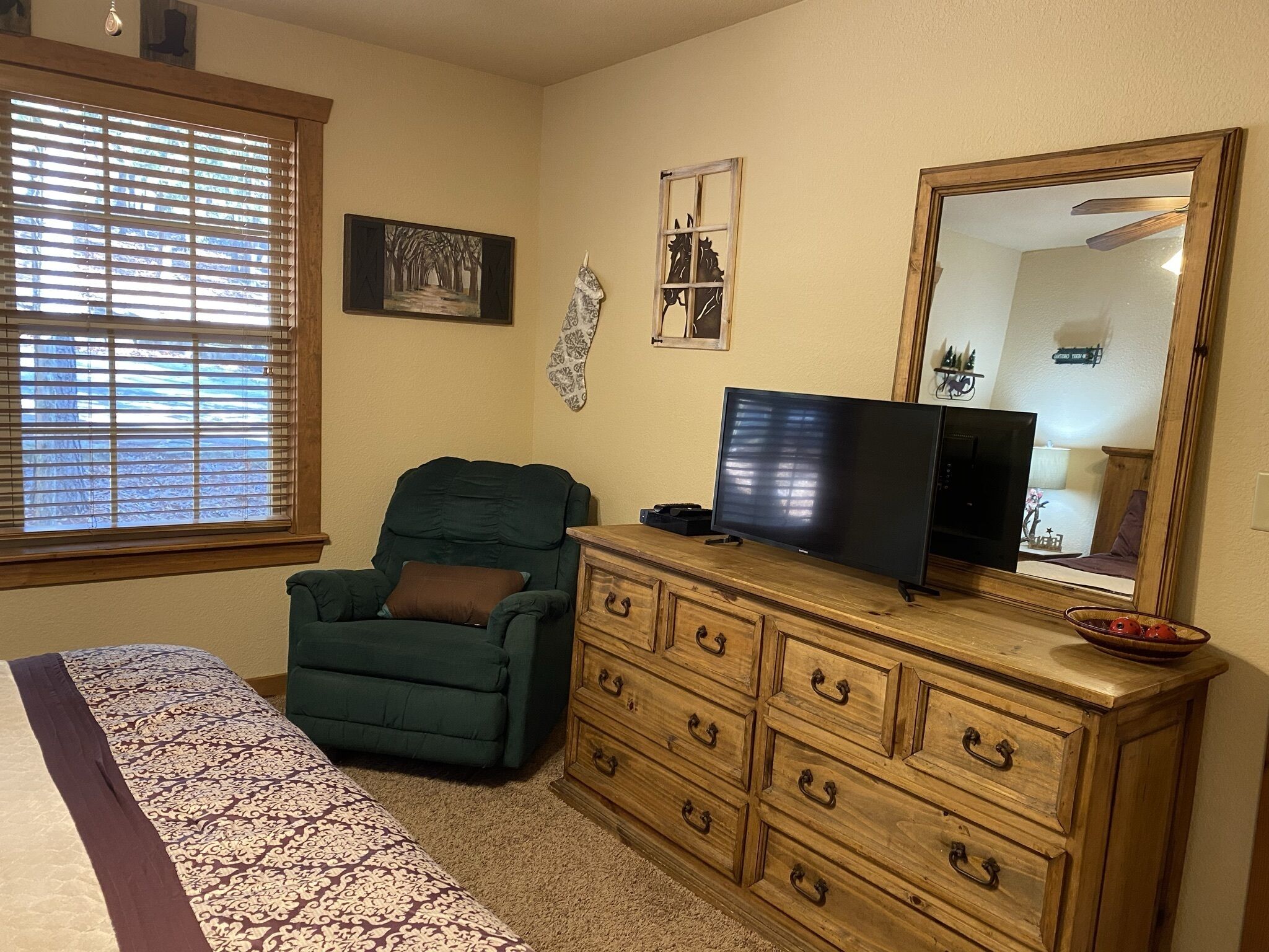Una habitación con una cama, un sillón verde, un aparador de madera, un televisor y una ventana con persianas.