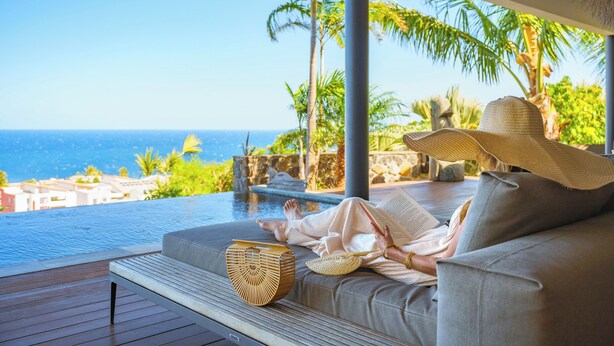A person relaxing on a sofa by a pool with a view of the ocean and palm trees. - Villa Coco Palm - Oasis de Luxe 5*- Magnificent Sea View, Private Pool & Jacuzzi (Saint-Pierre)