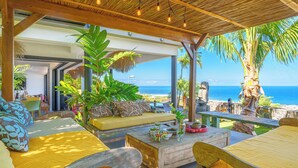A covered outdoor seating area with a view of the ocean, featuring wooden furniture, a thatched roof, and tropical plants. - Villa Coco Palm - Oasis de Luxe 5*- Magnificent Sea View, Private Pool & Jacuzzi (Saint-Pierre)