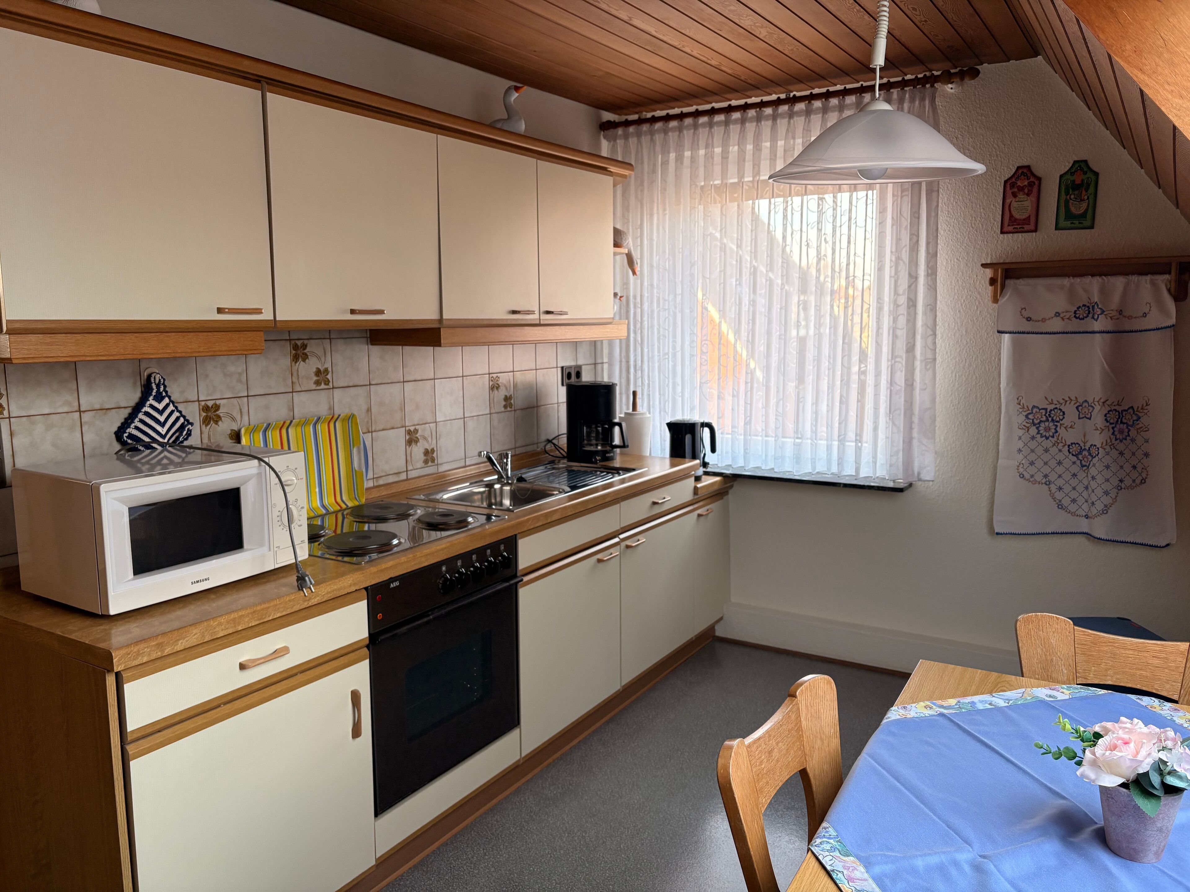 Een kleine keuken met witte kastjes, een magnetron, fornuis en spoelbak. Er is een raam met dunne gordijnen en een tafel met stoelen.