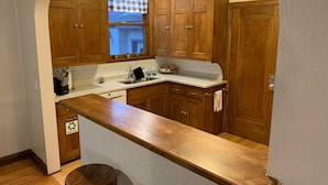 Eine Küche mit Holzausstattung, einer weißen Arbeitsplatte, einem Fenster mit karierter Gardine und einem Recyclingbehälter.